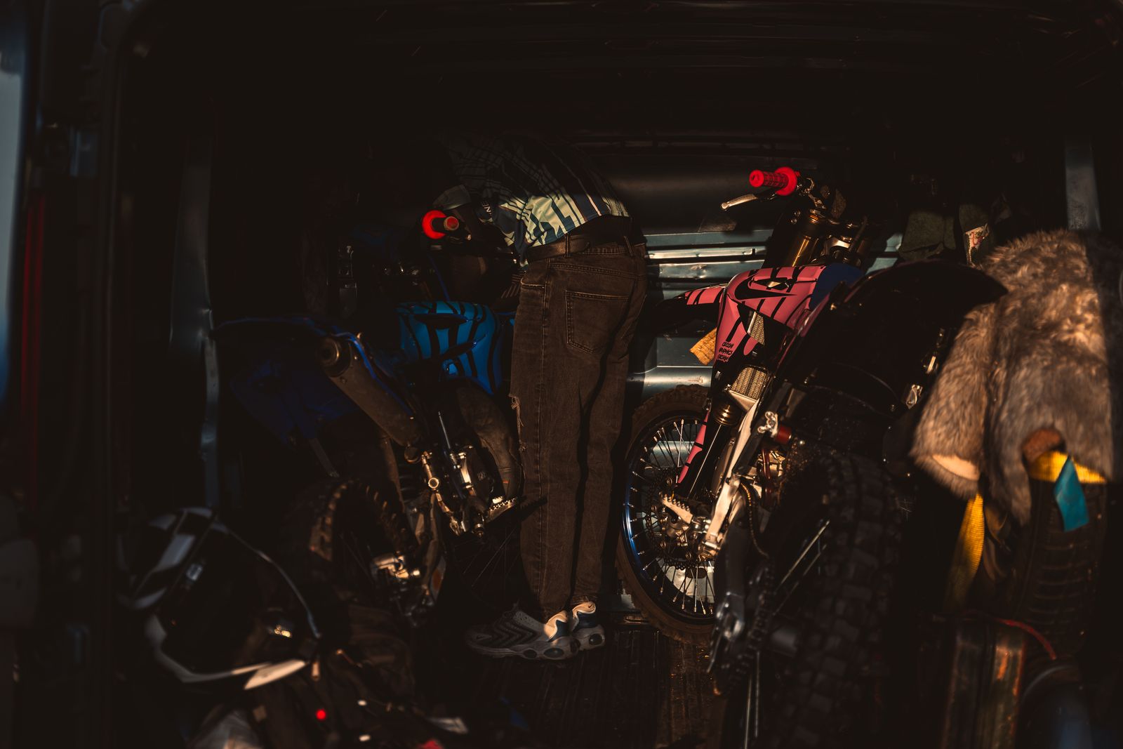 A rider loading blue and pink dirt bikes into the back of a van in low golden light