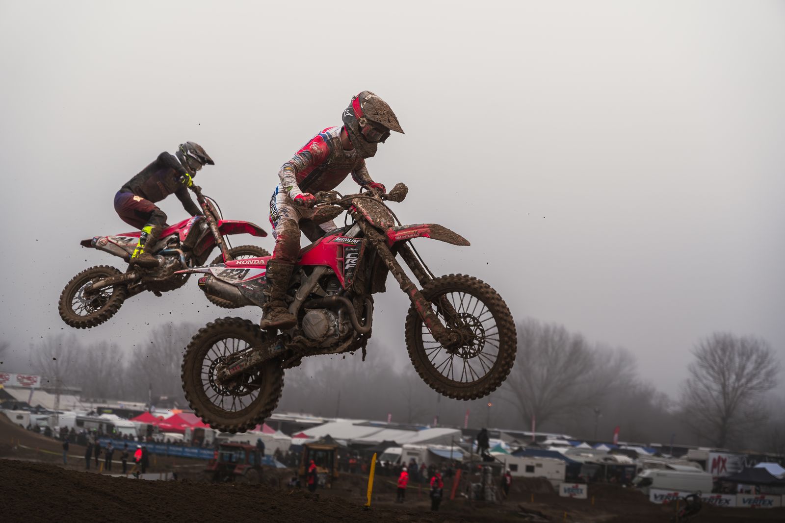 Two mud-covered Honda motocross bikes in mid-air over a jump on a foggy day