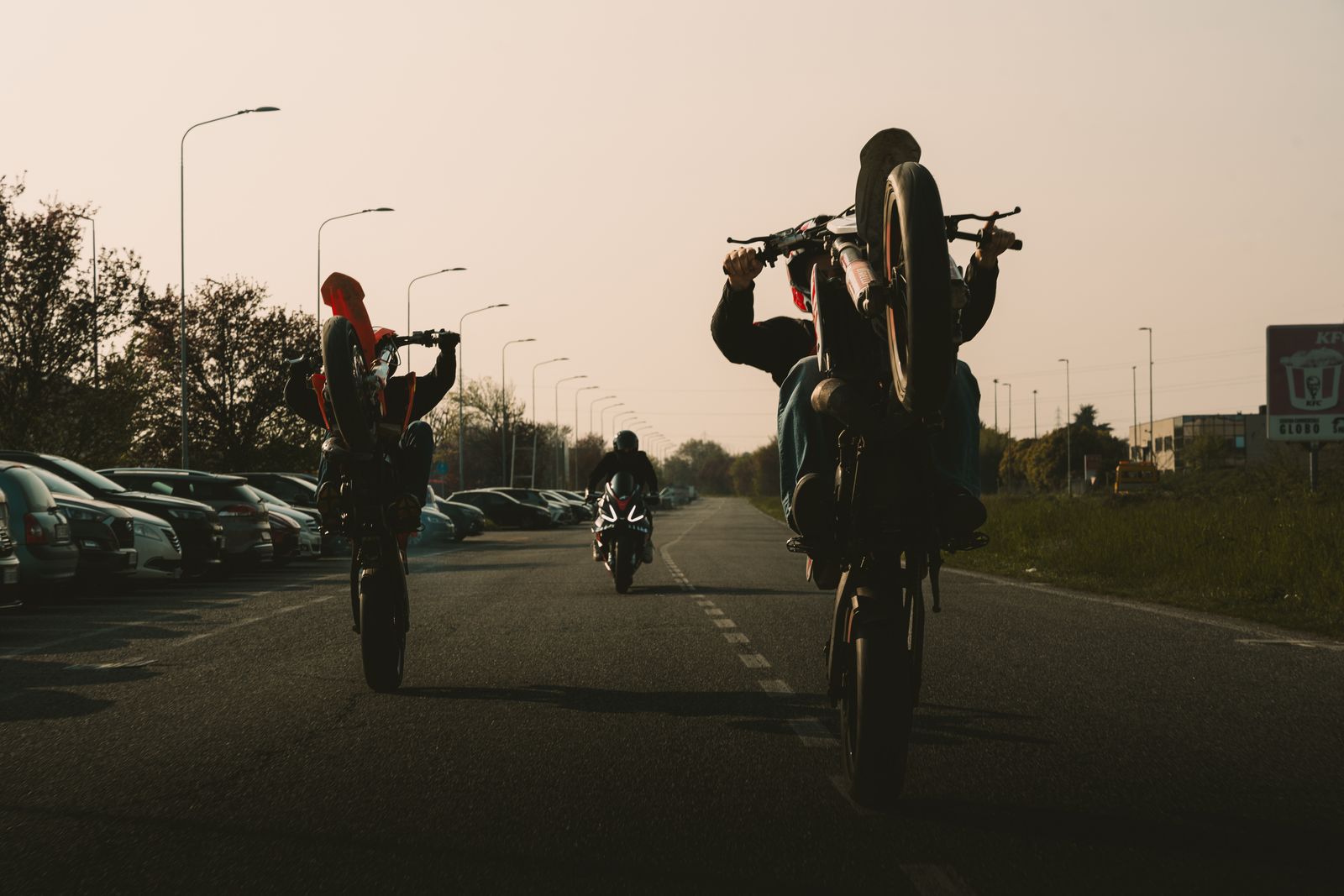 Two riders pulling wheelies side by side on a straight road at sunset, a sport bike following behind