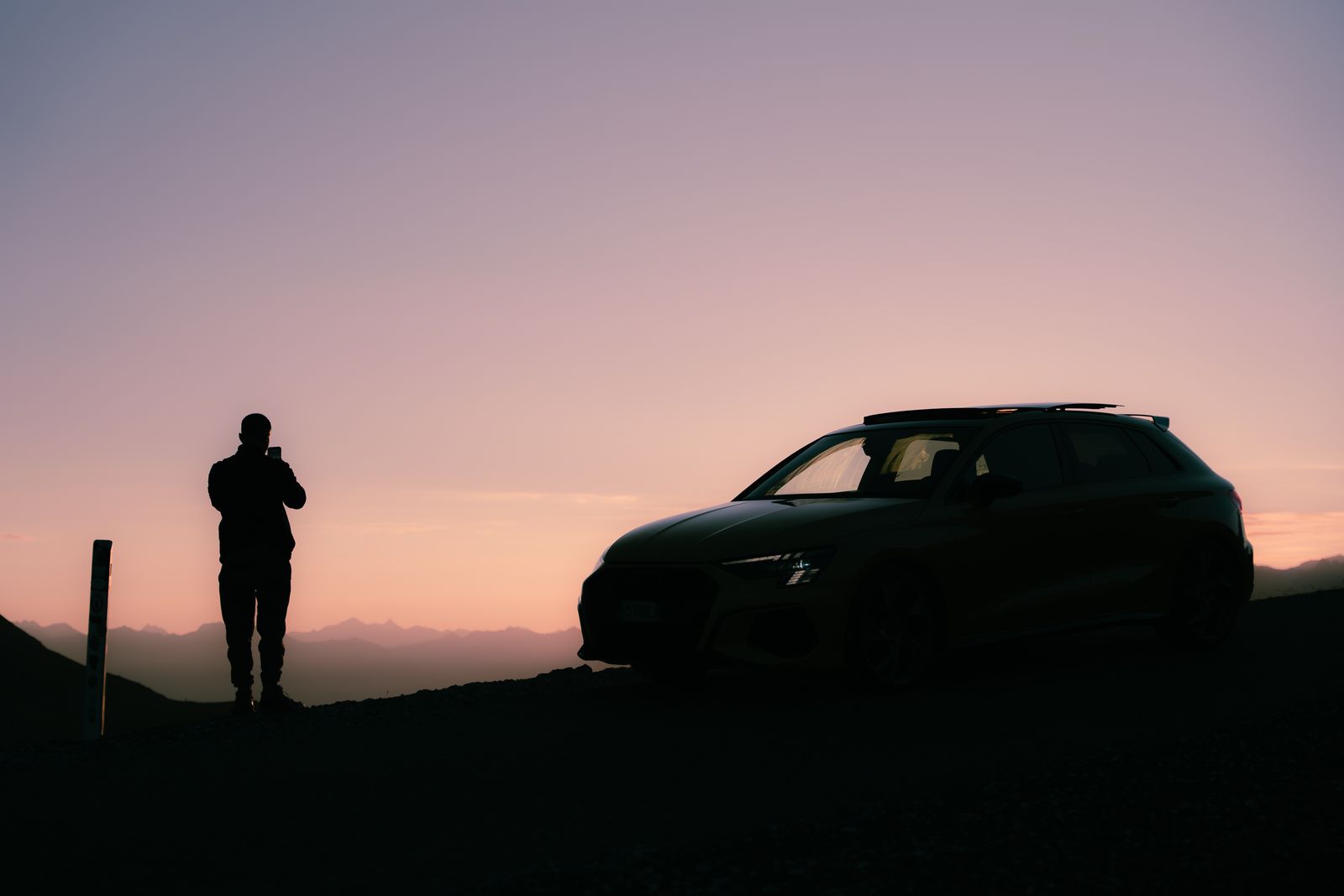 The silhouette of an Audi S3 ABT and a person taking a photo, against a pink sunrise sky over the mountains
