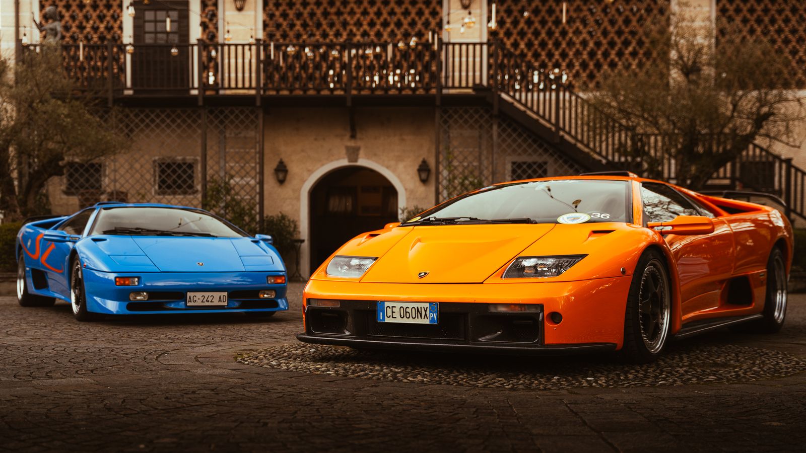 A blue Lamborghini Diablo SV and an orange Diablo GT parked side by side on cobbles in front of an old villa
