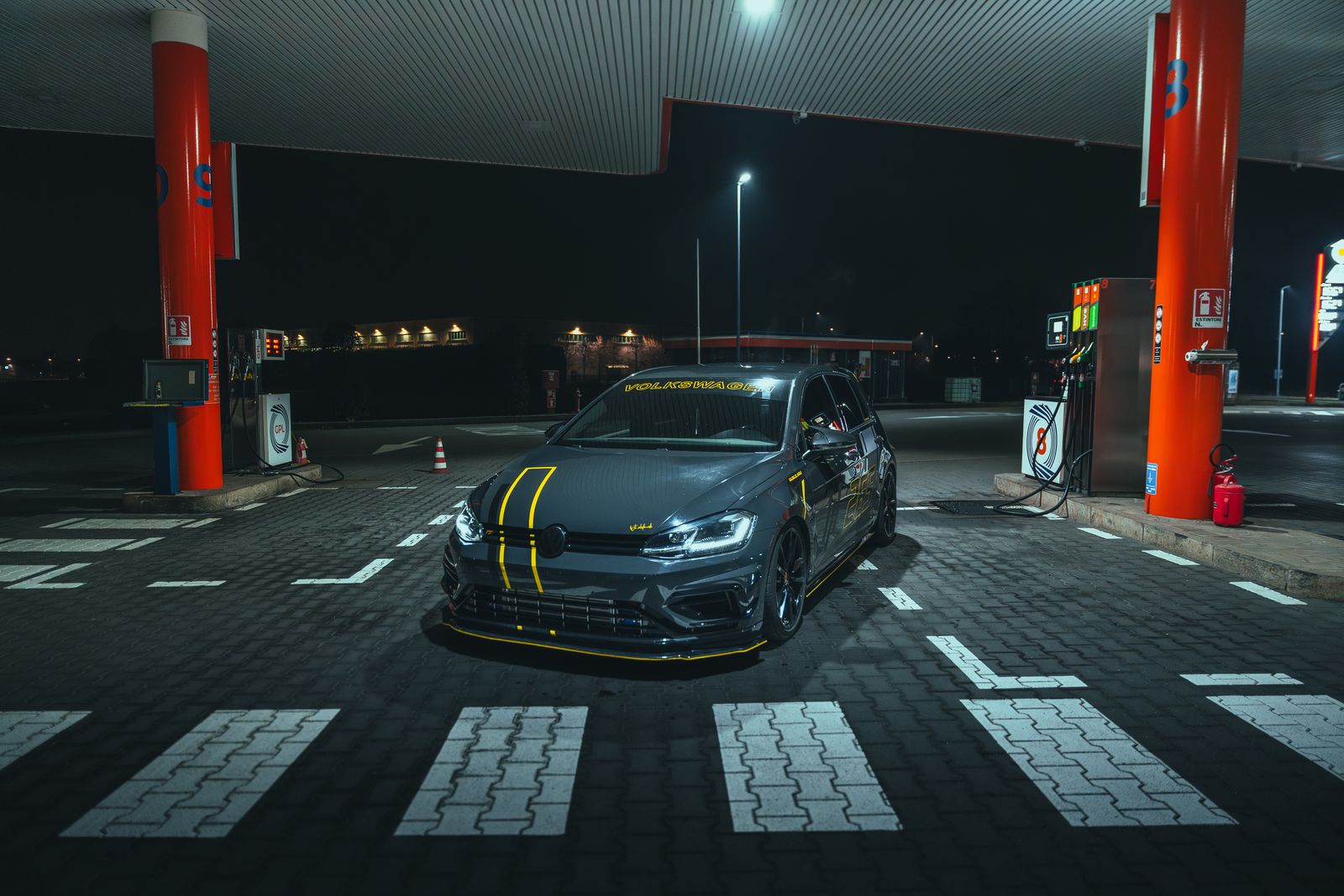 A grey Volkswagen Golf R with yellow stripes under the canopy of a petrol station at night, red pillars either side
