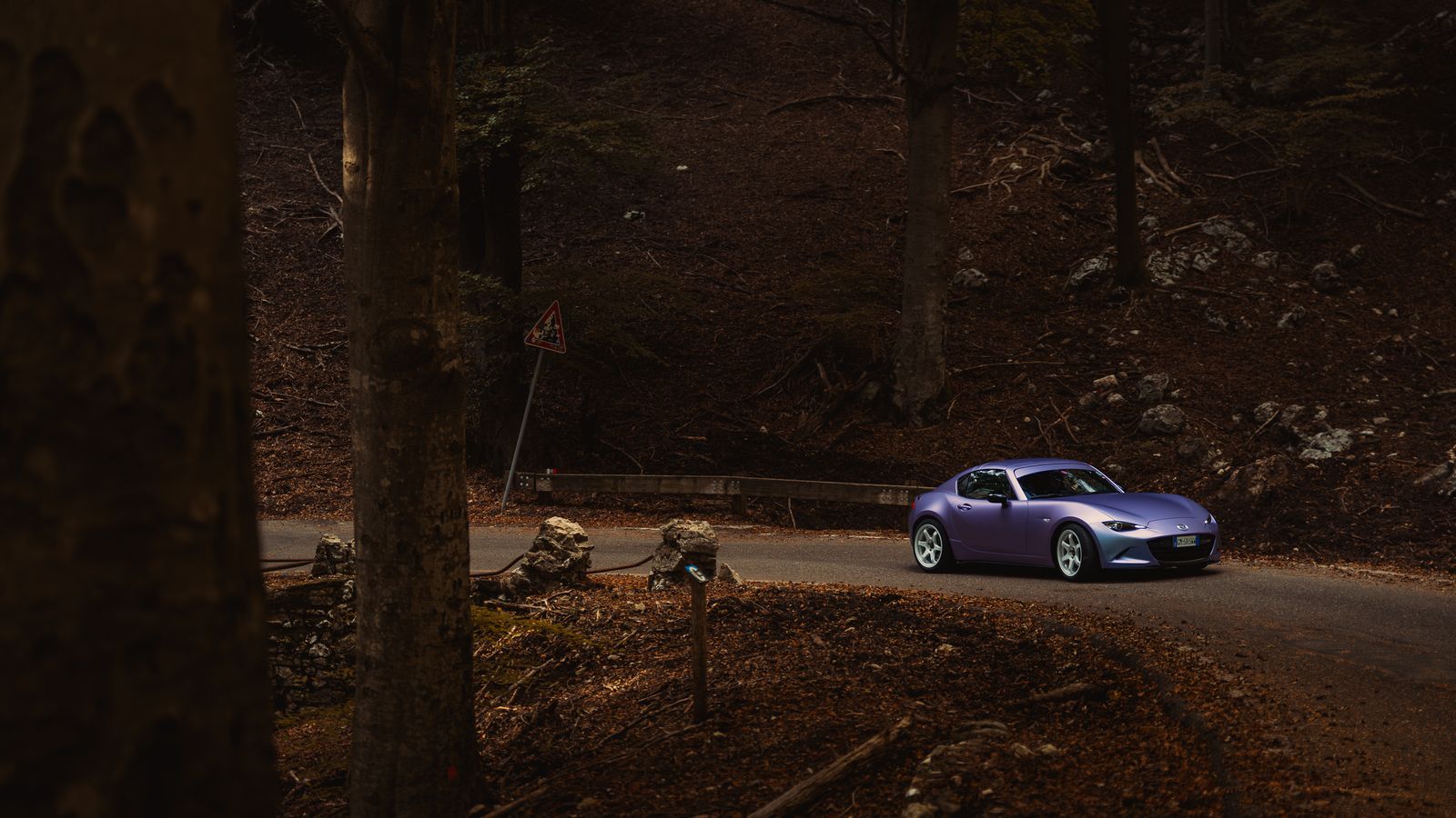 A matte lilac Mazda MX-5 RF taking a hairpin on a narrow road through a dark autumn forest