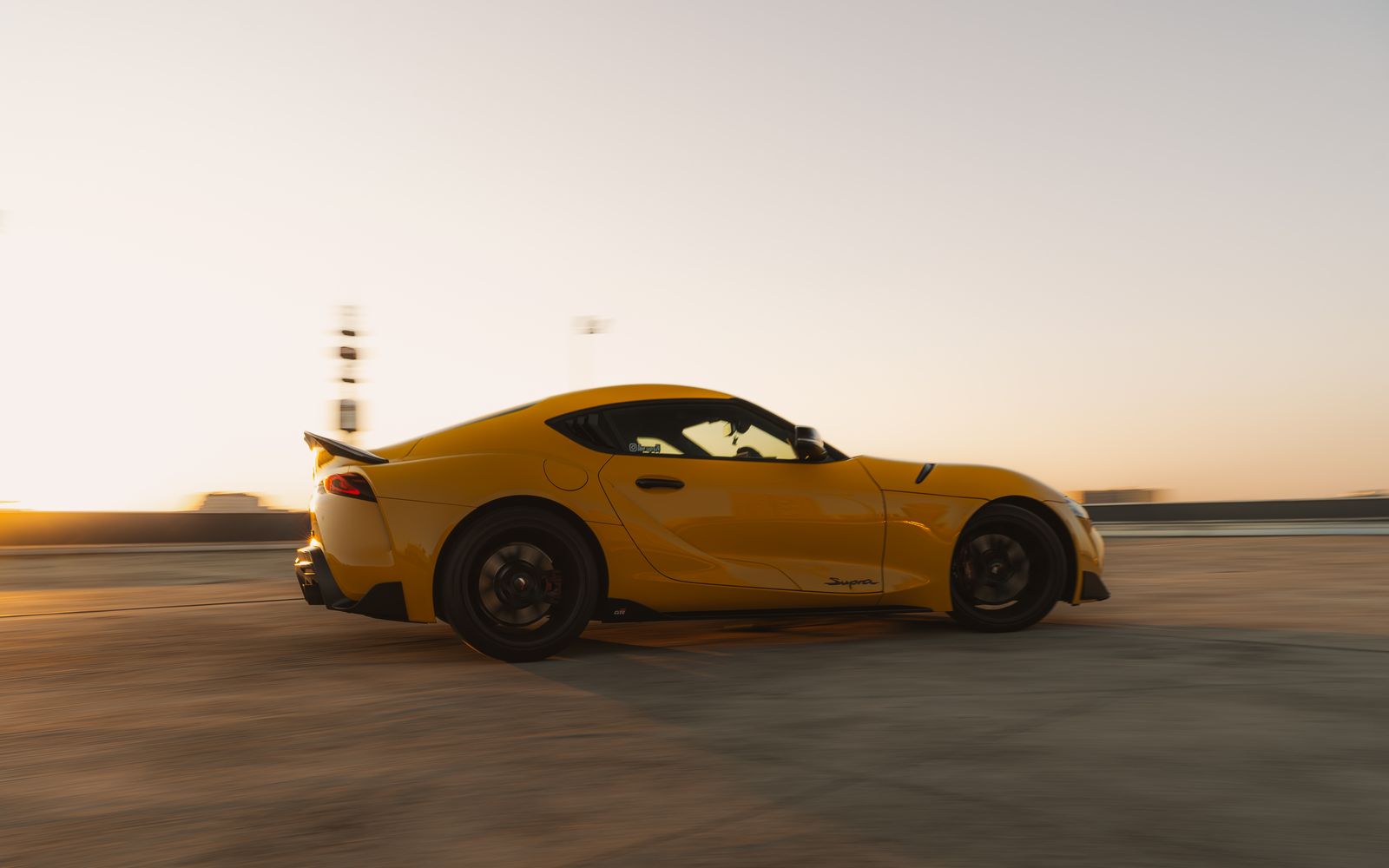 A yellow Toyota GR Supra in profile, moving, with the ground streaked and the low sun behind it