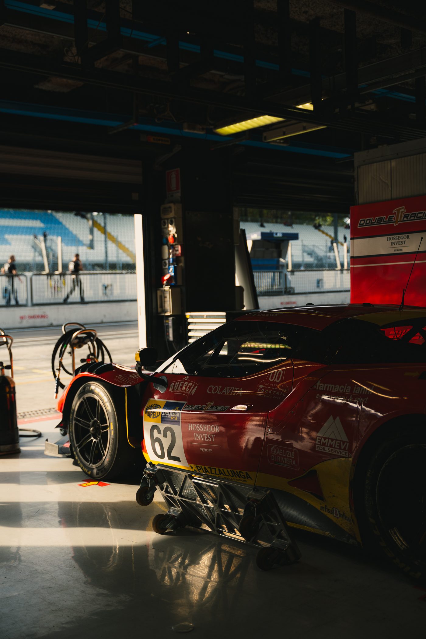 A red and yellow Ferrari race car, number 62, on its jacks in a pit garage, the empty grandstand beyond the pit lane