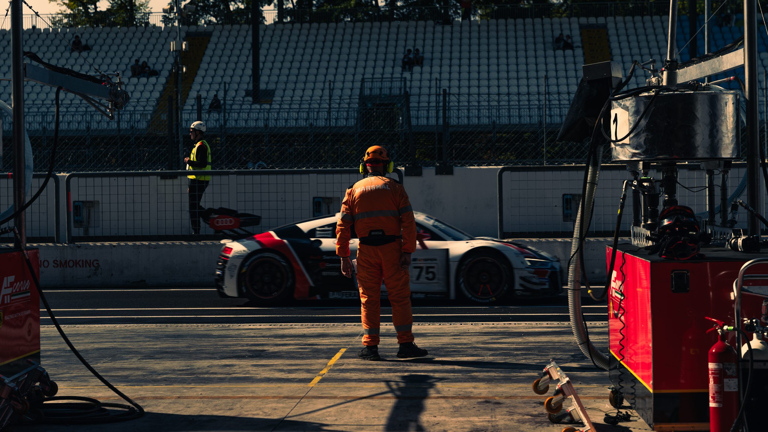 A marshal in orange overalls stands in the pit lane as a white Audi R8, number 75, passes in a blur, an empty grandstand behind