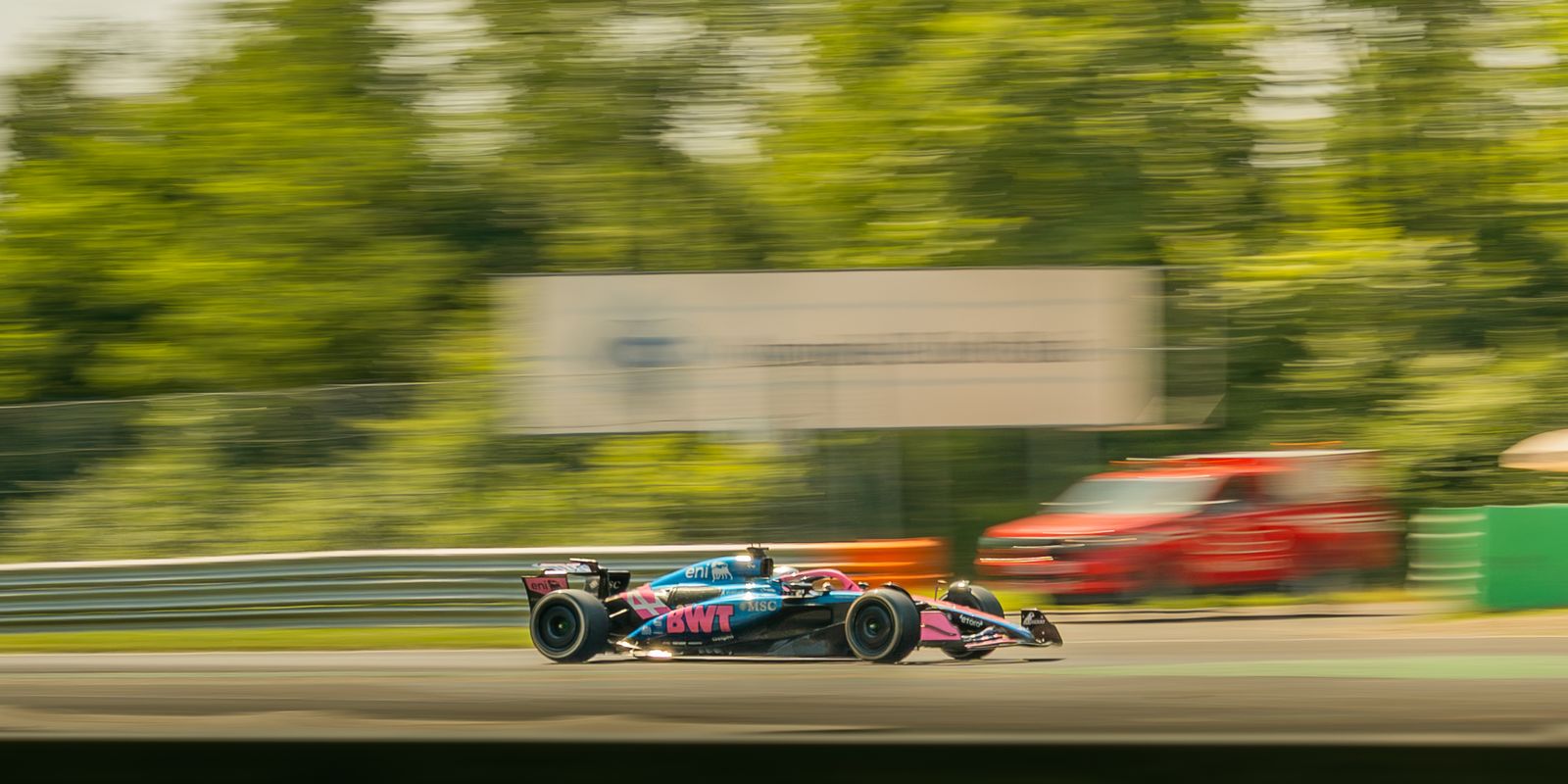 A blue and pink Alpine Formula 1 car panned along the straight, trees and a marshal car blurred behind