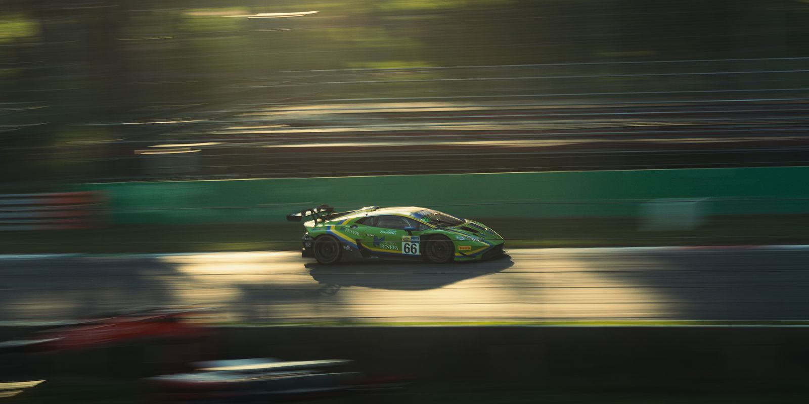 A green Lamborghini Huracán race car, number 66, panned through low golden sunlight with the barriers streaked behind