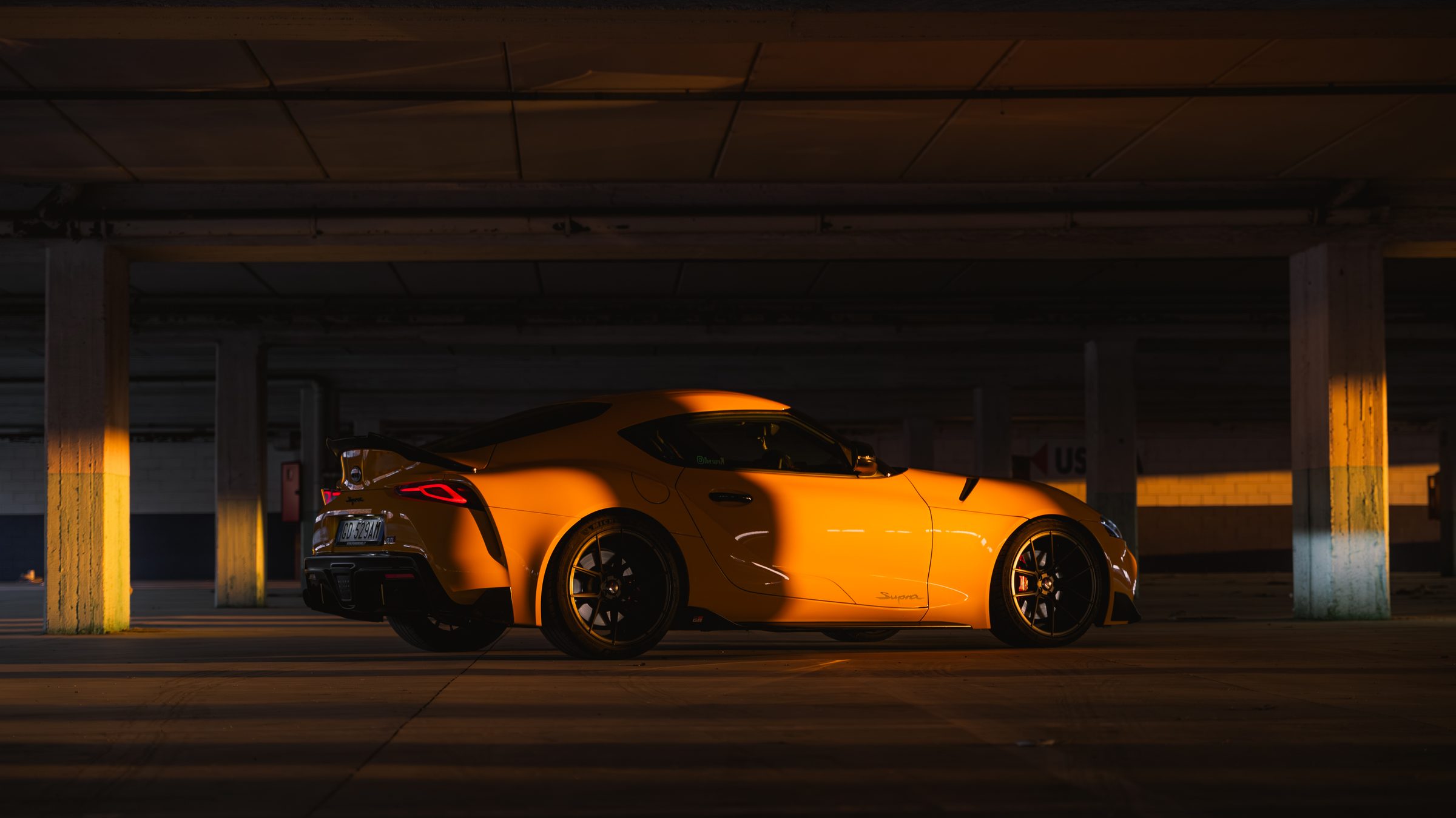A yellow Toyota GR Supra in profile in a dark car park, low golden sunlight falling across the car and the concrete pillars