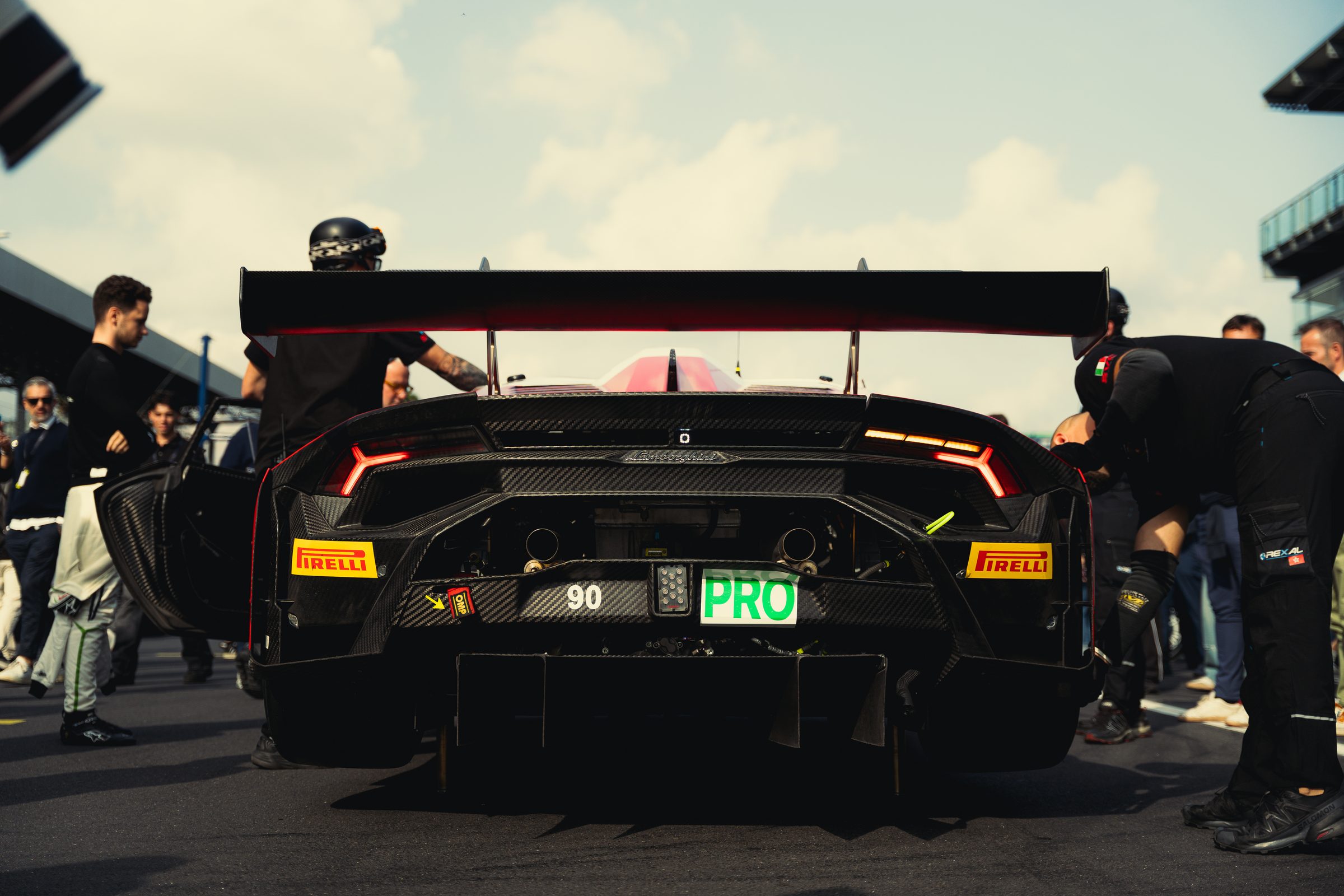 The carbon rear of a Lamborghini Huracán GT3, number 90, on the starting grid with mechanics working around it